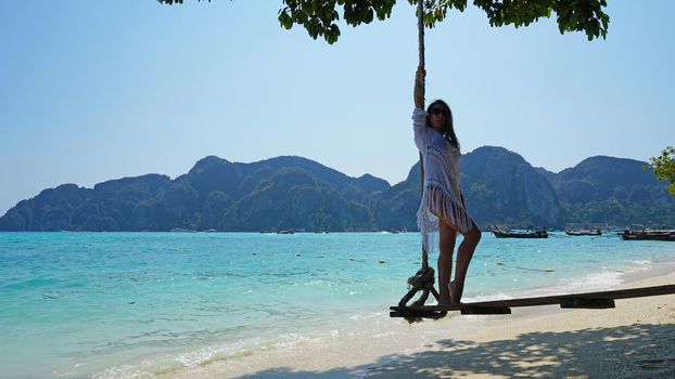 Beautiful girl in a white Cape and a black bathing suit on a swing. Swing on the beach. View of the island, beach with sand, blue water and green leaves of trees. Visible hills of the island. Thailand