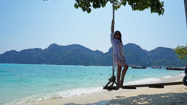 Beautiful girl in a white Cape and a black bathing suit on a swing. Swing on the beach. View of the island, beach with sand, blue water and green leaves of trees. Visible hills of the island. Thailand