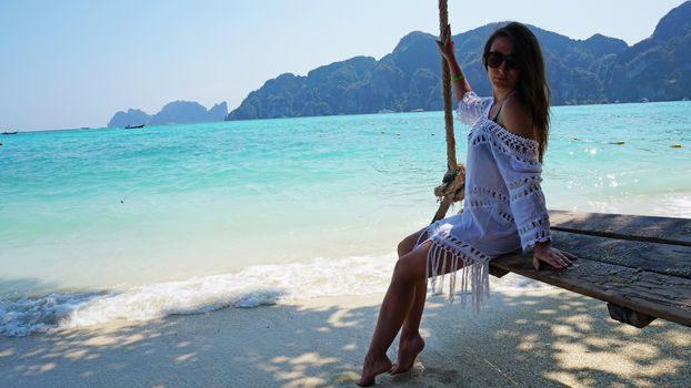 Beautiful girl in a white Cape and a black bathing suit on a swing. Swing on the beach. View of the island, beach with sand, blue water and green leaves of trees. Visible hills of the island. Thailand