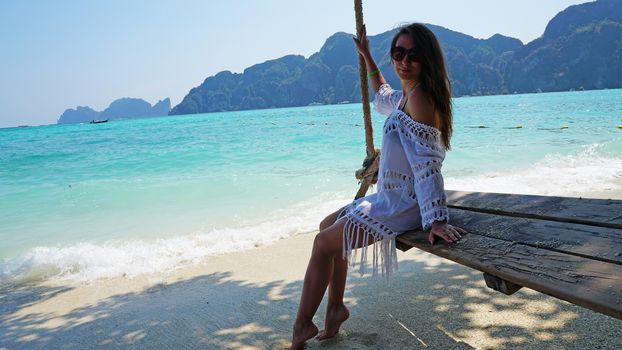 Beautiful girl in a white Cape and a black bathing suit on a swing. Swing on the beach. View of the island, beach with sand, blue water and green leaves of trees. Visible hills of the island. Thailand