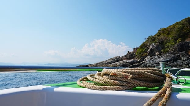 View of the bow of a fast boat with a rope. The metal rim glitters. It offers a landscape on the blue sea, a green island with trees and large stones. In the distance the blue sky with white clouds.