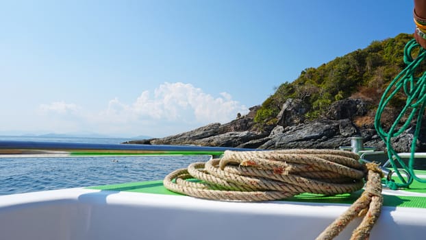 View of the bow of a fast boat with a rope. The metal rim glitters. It offers a landscape on the blue sea, a green island with trees and large stones. In the distance the blue sky with white clouds.