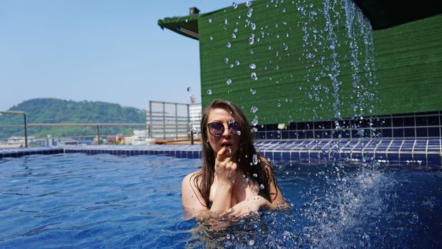 A girl in a black swimsuit and glasses poses on the edge of the pool. Swimming pool on the roof of the building. Photosession. View of the blue sky and clear water. Small waterfall. Thailand, Phuket.