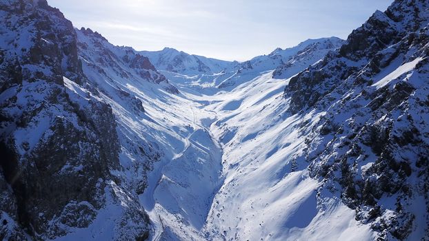 High snowy mountains, spruce trees grow in places. Huge rocks and cliffs, gorges where there may be an avalanche. Top view from a drone. Sunny day, shadows from the mountains fall on the snow. Almaty