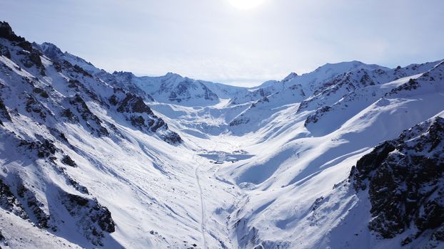 High snowy mountains, spruce trees grow in places. Huge rocks and cliffs, gorges where there may be an avalanche. Top view from a drone. Sunny day, shadows from the mountains fall on the snow. Almaty