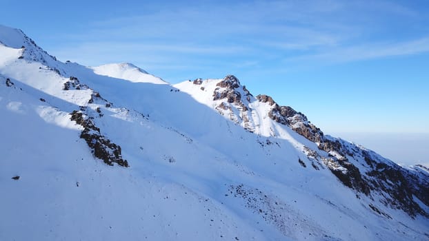 Snow mountain peaks with rocks. View from drone. In places, you can see glaciers, large rocks and rocks. Huge drifts of snow covered mountains. Dangerous place. Blue sky and sun. Shadow of the peaks.