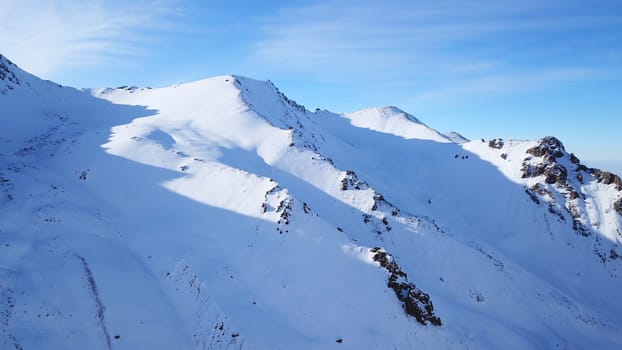 Snow mountain peaks with rocks. View from drone. In places, you can see glaciers, large rocks and rocks. Huge drifts of snow covered mountains. Dangerous place. Blue sky and sun. Shadow of the peaks.