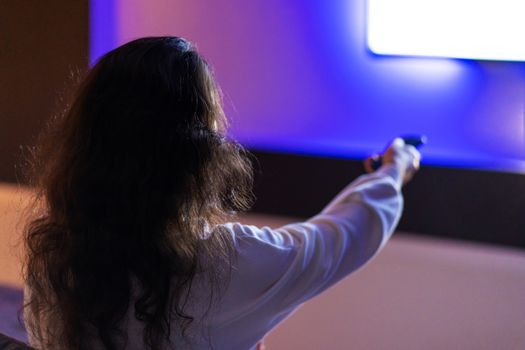 A person watches TV with a remote control in hand, a view from the back.