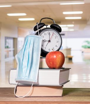 Concept for back to school with coronavirus or Covid-19 with books, alarm clock and apple with face mask against background of school corridor