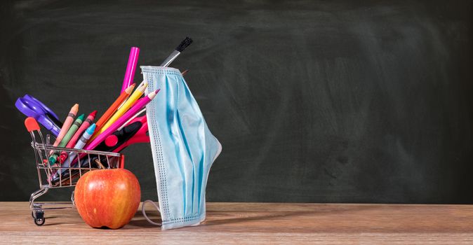 Back to school during coronavirus pandemic with pencils, crayons and face mask in shopping cart with apple against chalkboard