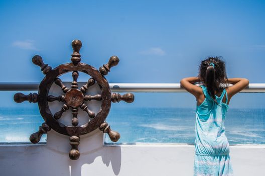 Girl observing the sea from a balcony of a house on the beach with a pirate ship helm at her side, enjoying life and summer vacation, rear view