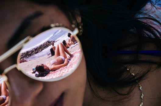 The glass of some sunglasses on a young woman reflects another woman on the sand with the background of the sea. Use it for a beach or summer concept