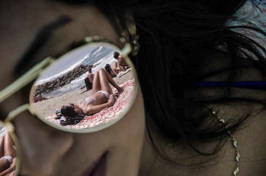 The glass of some sunglasses on a young woman reflects another woman on the sand with the background of the sea. Use it for a beach or summer concept