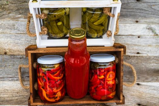 Wooden crate with bottles with tomatoes sauce and glass jars with pickled red bell peppers isolated in a rustic composition. Jars with variety of pickled vegetables preserved food concept.