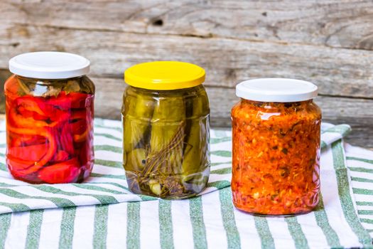 Glass jars with pickled red bell peppers and pickled cucumbers (pickles) isolated. Jars with variety of pickled vegetables. Preserved food concept in a rustic composition.