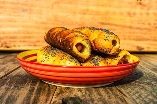 Sausages baked in dough sprinkled with salt and poppy seeds in a colorful plate. Sausages rolls, delicious homemade pastries in a rustic composition.