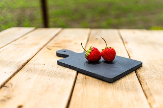 Close up of two strawberries on small black cutting board isolated outdoor on wooden table.