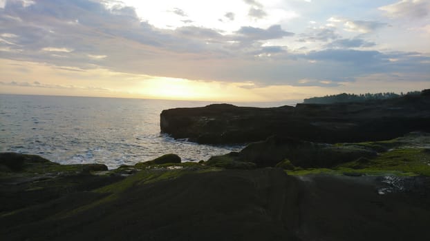 Sunset over the ocean. View of large waves breaking on the rocks. Clouds hang over the horizon. A ray from the sun is reflected on the water in orange color. The coast of Bali is visible.