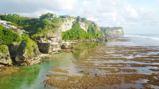 Beautiful beach among the rocks of the island of Bali. Uluwatu-surfers beach. Huge cliffs, greenery grows on the rocks, there are hotels and you can see big waves with clouds. But the tide is out.