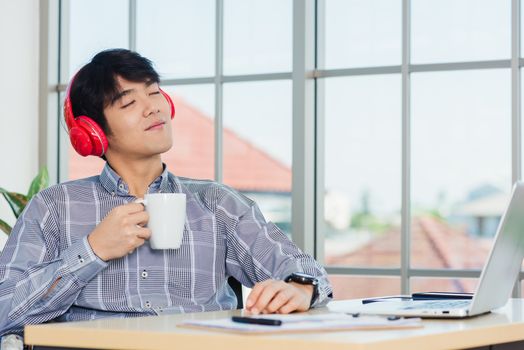 Asian young businessman happiness with red headphones sitting on desk workplace home office with a laptop computer, confident handsome man lifestyle smile relax holding a coffee cup, listens to music