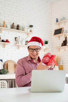 Christmas online greetings. Happy young man in santa hat greeting his friends in video chat or call on laptop sitting in his kitchen