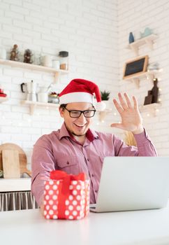 Christmas online greetings. Happy young man in santa hat greeting his friends in video chat or call on laptop sitting in his kitchen
