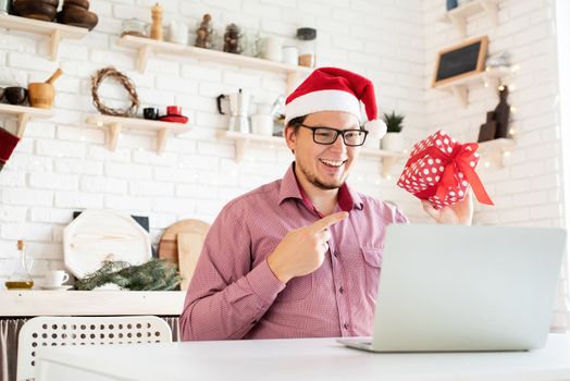 Christmas online greetings. Happy young man in santa hat greeting his friends in video chat or call on laptop sitting in his kitchen with copy space
