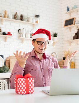 Christmas online greetings. Happy young man in santa hat greeting his friends in video chat or call on laptop sitting in his kitchen