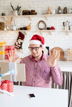 Christmas online greetings. Happy young man in santa hat greeting his friends in video chat or call on tablet sitting in his kitchen