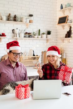 Christmas online greetings. Happy young couple in santa hats greeting their friends in a video call on laptop sitting at the kitchen