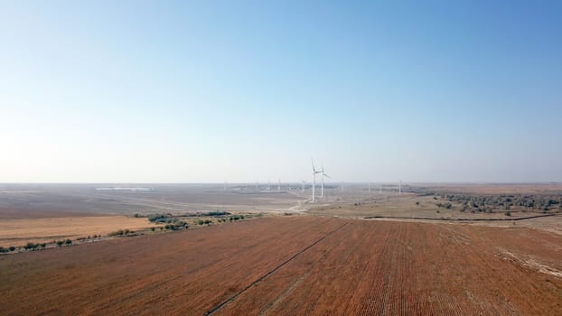 Windmills near the field. Top view from a drone. Blue sky, light shroud, windmills turn, green energy is generated. Alternative energy, clean land. Yellow plants, and a desert. Industrial farm.