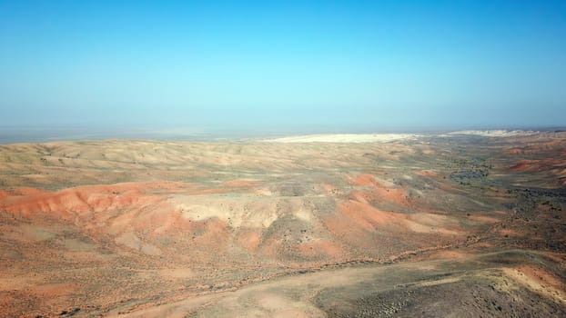 Colored hills of the gorge in the desert. Top view from drone of the red-orange-yellow hills. Cut in different layers, like epochs. Clay of the mountain. Low dry bushes, cracks, blue sky. Kazakhstan.