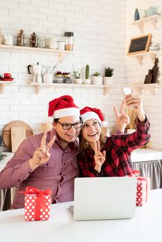 Christmas online greetings. Happy young couple in santa hats greeting their friends in a video call on laptop and mobile phone sitting at the kitchen