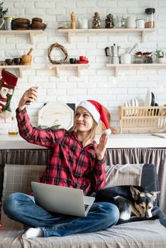 Chhristmas online greetings. Happy young woman in santa hat greeting her friends in video call on laptop and mobile phone