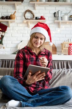 Christmas online greetings. young blond woman in santa hat working on tablet sitting on the couch