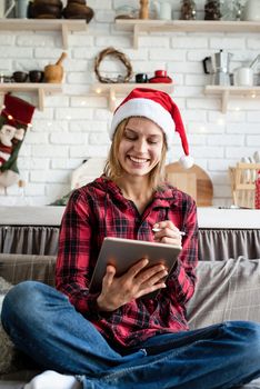 Christmas online greetings. young blond woman in santa hat working on tablet sitting on the couch