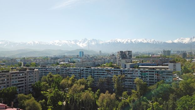 View of Almaty city with white clouds and blue sky. The green city is completely covered with trees, clean roads, transport, houses. On the hill stands the Kok Tobe TV tower. Mountains in the distance