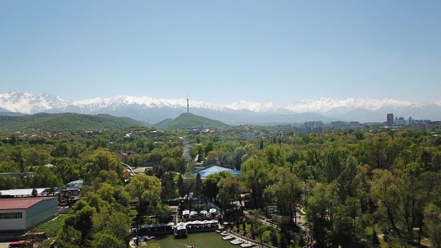 Green city Park with a pond and views of the mountains of the TRANS-ili Alatau. Lots of green trees, blue sky, snowy mountains and the Kok Tobe TV tower. Green water in the pond. Roofs are visible.