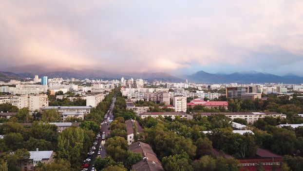 Bright color sunset over the city of Almaty. Huge clouds over the mountains and the city shimmer from bright blue to yellow and dark blue. Tall houses and green trees, cars driving on the roads.