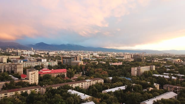 Bright color sunset over the city of Almaty. Huge clouds over the mountains and the city shimmer from bright blue to yellow and dark blue. Tall houses and green trees, cars driving on the roads.