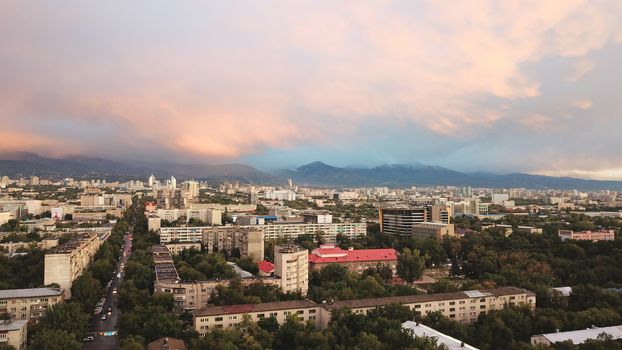 Bright color sunset over the city of Almaty. Huge clouds over the mountains and the city shimmer from bright blue to yellow and dark blue. Tall houses and green trees, cars driving on the roads.