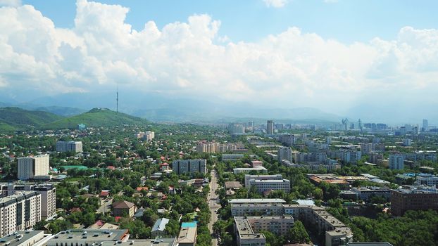 View of Almaty city with white clouds and blue sky. The green city is completely covered with trees, clean roads, transport, houses. On the hill stands the Kok Tobe TV tower. Mountains in the distance