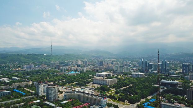 View of Almaty city with white clouds and blue sky. The green city is completely covered with trees, clean roads, transport, houses. On the hill stands the Kok Tobe TV tower. Mountains in the distance