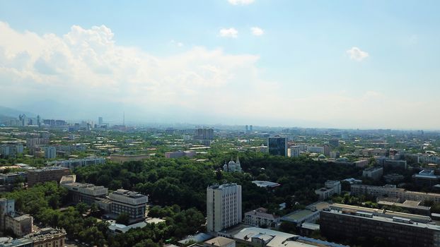 View of Almaty city with white clouds and blue sky. The green city is completely covered with trees, clean roads, transport, houses. On the hill stands the Kok Tobe TV tower. Mountains in the distance