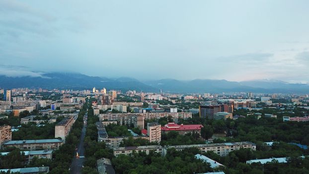 Clouds over the mountains and the city of Almaty at sunset. A lot of green trees, cars driving on the road, high mountains can be seen in the distance. On the hill stands the Kok Tobe TV tower.