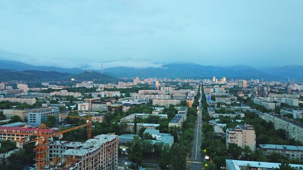 Clouds over the mountains and the city of Almaty at sunset. A lot of green trees, cars driving on the road, high mountains can be seen in the distance. On the hill stands the Kok Tobe TV tower.