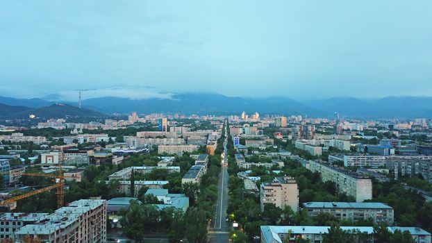 Clouds over the mountains and the city of Almaty at sunset. A lot of green trees, cars driving on the road, high mountains can be seen in the distance. On the hill stands the Kok Tobe TV tower.