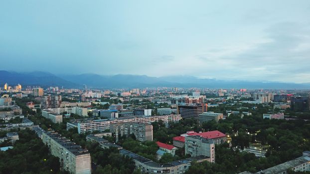 Clouds over the mountains and the city of Almaty at sunset. A lot of green trees, cars driving on the road, high mountains can be seen in the distance. On the hill stands the Kok Tobe TV tower.