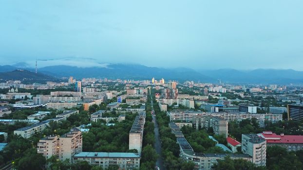 Clouds over the mountains and the city of Almaty at sunset. A lot of green trees, cars driving on the road, high mountains can be seen in the distance. On the hill stands the Kok Tobe TV tower.