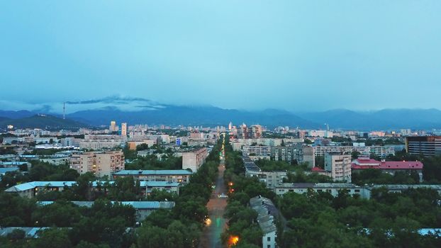 Clouds over the mountains and the city of Almaty at sunset. A lot of green trees, cars driving on the road, high mountains can be seen in the distance. On the hill stands the Kok Tobe TV tower.